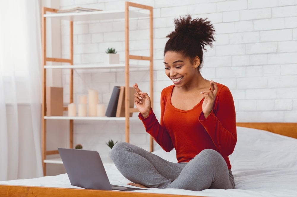 Excited lady sat in a natural colour bedroom on a bed with a laptop.